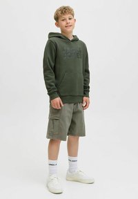 Zelena kapuca s prednjim žepom i crnim logotipom, uparena sa svijetložutim cargo kratkim hlačama, bijelim tenisicama i čarapama s logotipom. Neutralna pozadina.