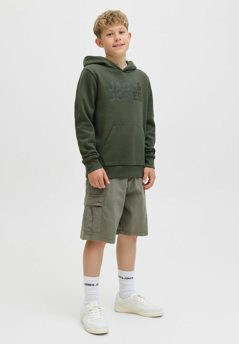 Zelena kapuca s prednjim žepom i crnim logotipom, uparena sa svijetložutim cargo kratkim hlačama, bijelim tenisicama i čarapama s logotipom. Neutralna pozadina.