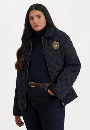 Black quilted jacket with a zip front, pointed collar, and emblem at the chest. Worn over a dark turtleneck and paired with dark jeans.