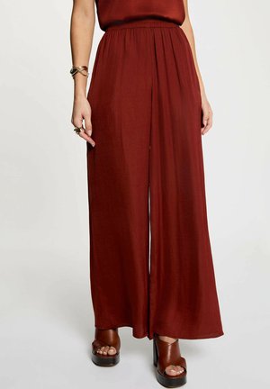 Donna con pantaloni ampi a vita alta colore ruggine, abbinati a sandali marroni con plateau e tacchi, e braccialetti e anelli d'oro alla mano sinistra.