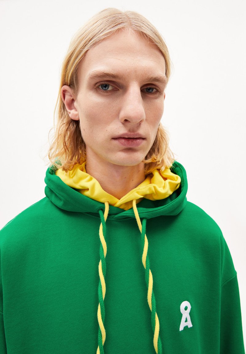 Grüne Kapuzenjacke aus weichem Stoff, mit einer gelben Kordel und einer inneren gelben Kapuze. Ein weißes Logo befindet sich auf der linken Seite.