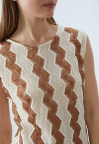 Strikket topp med et teksturert zigzag-mønster i brunt og krem, med rund hals og uten ermer.