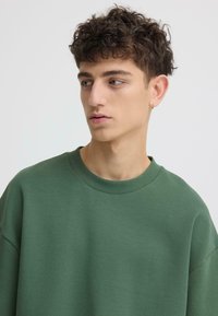 Sweat-shirt vert avec une coupe décontractée, encolure ronde et épaules légèrement tombantes. Texture lisse sans motifs ni accents visibles.