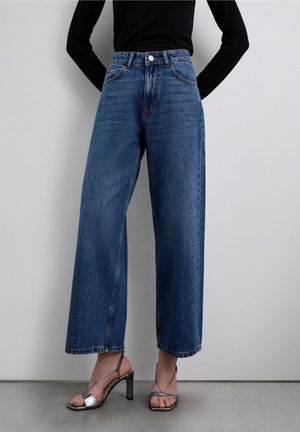 Femme portant un jean bleu taille haute à jambes larges avec de subtiles taches et des sandales argentées à talons hauts fines et à brides, debout contre un mur uni.