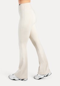 Lichtbeige uitlopende leggings gemaakt van zacht, rekbaar materiaal met een hoge tailleband en naaddetails, gecombineerd met witte sneakers.