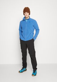 Jasnoniebieska polarowa kurtka z zamkiem, czarne spodnie i brązowa dzianinowa czapka. Jasnoniebieskie buty biegowe z czarnymi i złotymi akcentami. Prosty design, luźny krój.