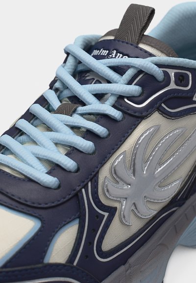 Κοντινό πλάνο αθλητικού παπουτσιού σε ναυτικό μπλε και μπεζ χρώμα με γαλάζια κορδόνια και ασημένιο αφηρημένο φύλλο λογότυπο στην πλευρική επιφάνεια.