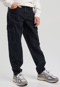 Pantaloni cargo in denim scuro con tasche laterali, vestibilità rilassata e cuciture a contrasto, abbinati a sneakers grigie e bianche.