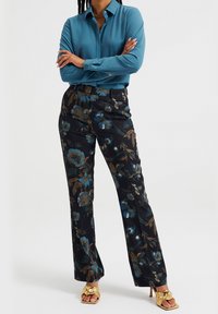 Teal skjorta med knappar i kombination med svarta blommiga byxor som har blå och bruna mönster; modell som bär metalliska guld klackar.