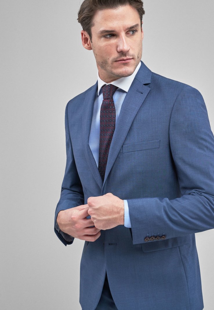 Chaqueta de traje ajustada de color azul claro en tejido texturizado con un solo bolsillo en el pecho, pantalones a juego y una corbata estampada con tonos oscuros.