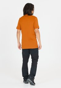 Herrtröja med kort ärm i orange, mjukt material och en figurnära design, kombinerad med svarta byxor med förstärkta detaljer och kraftiga stövlar.