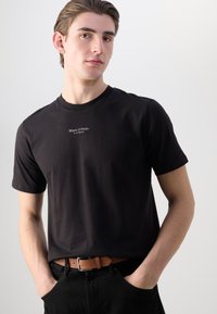 Tricou din bumbac negru cu guler rotund și mâneci scurte. Prezintă un logo de marcă imprimat alb la piept. Modelul poartă blugi negri și o curea maro.
