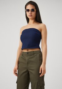 Tamsiai mėlynos džinsinės be rankovių palaidinė, prilipusi prie kūno, derinama su alyvuogių spalvos krovinių kelnėmis, turinčios sagčių užsegimą ir šonines kišenes.