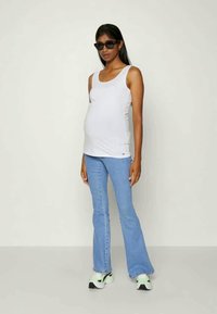 Top justo branco, jeans flare azul claro e óculos de sol pretos. Ténis com detalhes verdes e sola preta. Modelo exibe uma postura relaxada.