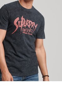 Camiseta gráfica negra con un acabado desgastado, que presenta letras rojas para "Superdry" y "Japan Tour". Cuello redondo clásico y mangas cortas.