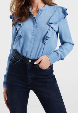 Hellblaue Jeansbluse mit Rüschenakzenten an den Schultern und der Knopfleiste, kombiniert mit dunkelblauen Jeans und einem goldenen Knopf.
