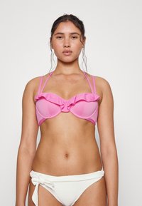Top de bikini estruturado em rosa com decote franzido, alças ajustáveis e aros para suporte. Combinado com parte de baixo de bikini branca com textura e nó.
