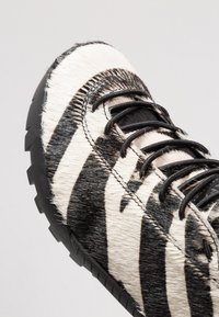 Calzado de rayas blancas y negras con un material texturizado similar a la piel de animal, punta redondeada y cordones negros sobre una suela de goma con detalles de tracción.