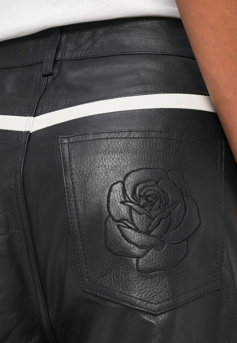 Pantalones de cuero negros con una franja horizontal blanca y un diseño de rosa en relieve en el bolsillo trasero, combinados con un top blanco de canalé.