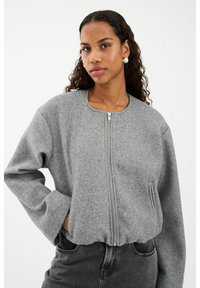 Kobieta z kręconymi włosami, nosząca srebrne kolczyki w kształcie kół, szary dres z zamkiem błyskawicznym oraz ciemnoszare dżinsy, stojąca z jedną ręką w kieszeni.