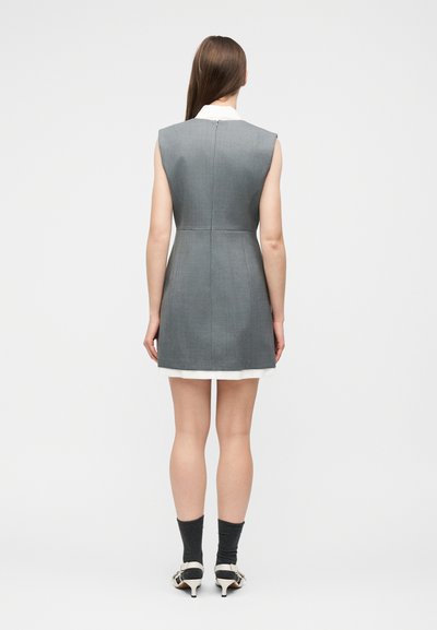 Γυναίκα που στέκεται με την πλάτη, φοράει μίνι αμάνικο γκρι φόρεμα με λευκό γιακά, μαύρες κάλτσες και λευκά παπούτσια με τακούνι σε απλό φόντο.