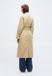 Femme portant un trench-coat beige ceinturé et un jean large bleu, debout de dos devant un fond blanc uni.