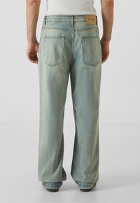 Světle modré široké džíny vyrobené z denimu, s dvěma zadními kapsami a koženou značkovou etiketou na opasku. Hladká textura.