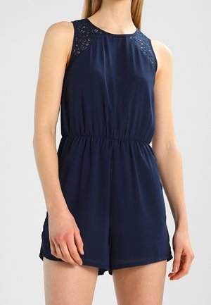 Ärmelloser marineblauer Overall mit elastischem Bund und Spitzen-Schultereinsätzen, getragen von einer stehenden Person vor einem einfarbigen Hintergrund.