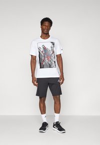 Hvit T-skjorte med Spider-Man grafikk, svarte shorts, hvite sokker og svarte joggesko. T-skjorten har rund hals og korte ermer.