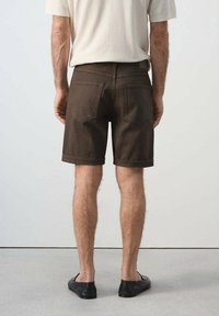 Hombre con pantalones cortos marrón oscuro hasta la rodilla, camisa blanca de manga corta y zapatos negros sin cordones, de espaldas sobre un fondo liso.