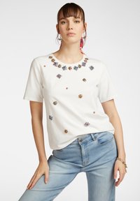 T-shirt bianca in cotone con maniche corte, scollatura rotonda e decorazioni floreali sparse in blu, verde e viola sulla parte anteriore.