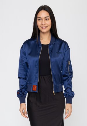 Kobieta ubrana w błyszczącą granatową kurtkę typu bomber na czarnym topie i spódnicy, stojąca na tle jasnoszarego, jednolitego tła.