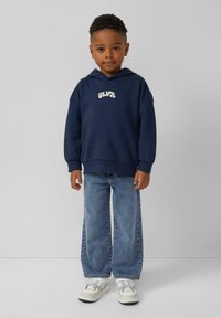 Granatowy hoodie z logo z przodu, zestawiony z jasnoniebieskimi szerokimi spodniami dżinsowymi oraz białymi trampkami z szarymi akcentami, prezentowany na neutralnym tle.