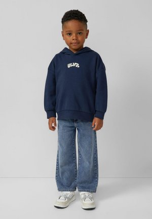 Marineblauwe hoodie met logo aan de voorkant, gecombineerd met lichtblauwe wijde jeans en witte sneakers met grijze accenten, weergegeven tegen een neutrale achtergrond.