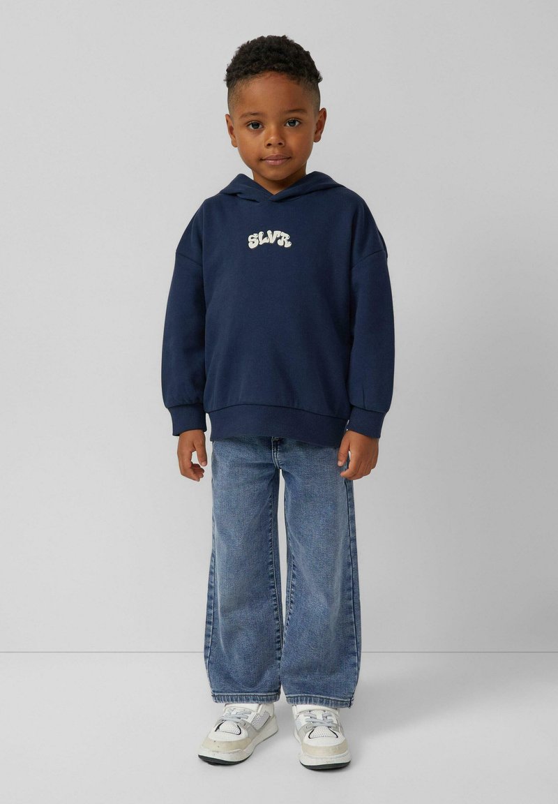 Granatowy hoodie z logo z przodu, zestawiony z jasnoniebieskimi szerokimi spodniami dżinsowymi oraz białymi trampkami z szarymi akcentami, prezentowany na neutralnym tle.