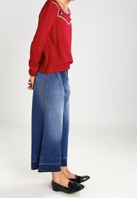Rode blouse met lange mouwen en een decoratieve halslijn, gecombineerd met brede blauwe spijkerbroek en zwarte instappers. Soepele stof en een relaxte pasvorm.