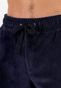Pantalones de pana en color azul marino con una textura acanalada, cinturilla elástica y cordón ajustable. Presentan ojales plateados para el sujeción del cordón.