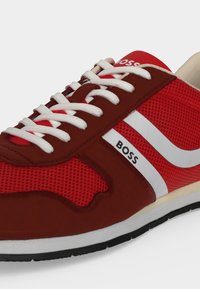 Sneaker homme rouge et blanc avec lacets blancs, marquage "BOSS" noir sur le côté et semelle noire avec dessus en tissu rouge texturé.