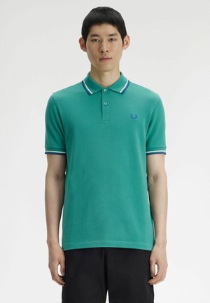Joven de pie, vistiendo una camiseta polo color azul verdoso con cuello y puños de manga blancos con rayas azules, y pantalones negros.