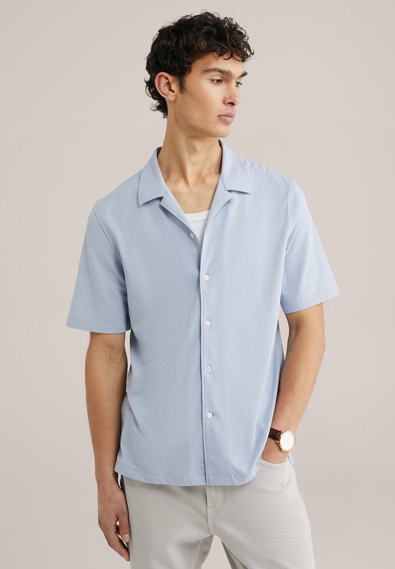 Camisetta azzurra a maniche corte con colletto rilassato e chiusura frontale con bottoni. Texture liscia, abbinata a pantaloni chiari e un orologio.
