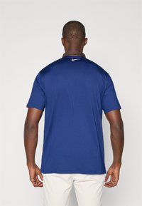 Una camiseta polo de manga corta en azul marino hecha de una tela ligera; cuenta con un collar marrón y un pequeño logo de Nike en blanco en la parte posterior cerca del cuello.