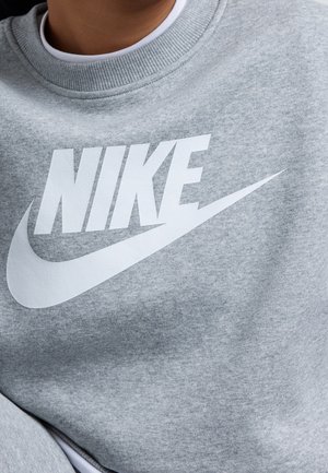Personne portant un sweat-shirt gris avec un grand logo Nike blanc et un swoosh sur la poitrine, superposé à un t-shirt blanc à col rond.