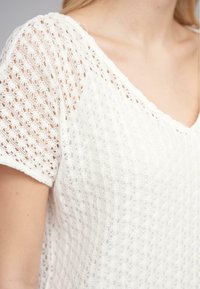 Hvit strikket topp med et blondeaktig mønster, med V-hals og kort erm. Stoffet er lett og semitransparent.