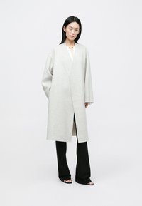 Manteau gris clair en mélange de laine, avec un design portefeuille, des manches larges et une coupe mi-longue, porté avec un pantalon noir et des sandales noires à lanières.