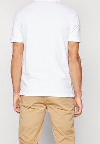 T-shirt blanc à manches courtes en coton ; col rond et fentes latérales à l'ourlet ; porté avec un pantalon kaki clair.