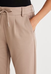 Beige kalhoty s páskem na utažení, sklady na přední straně a bočními kapsami. Tkanina vypadá jemně a hladce s dobře padnoucím střihem.