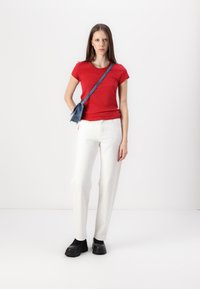 Röd åtsittande t-shirt, vita raka byxor och svarta kraftiga skor. Liten denimcrossbody-väska med en blå rem. Enfärgad bakgrund.