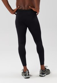 Juodos sportinės legginės su lygiu paviršiumi, turinčios platų juosmenį. Mūvimos su pilkais ir juodais bėgimo bateliais. Atsižvelgiama į užpakalinį vaizdą.