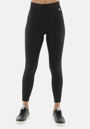 Person wearing black high-waisted leggings and black sneakers with white soles, standing against a plain light background.