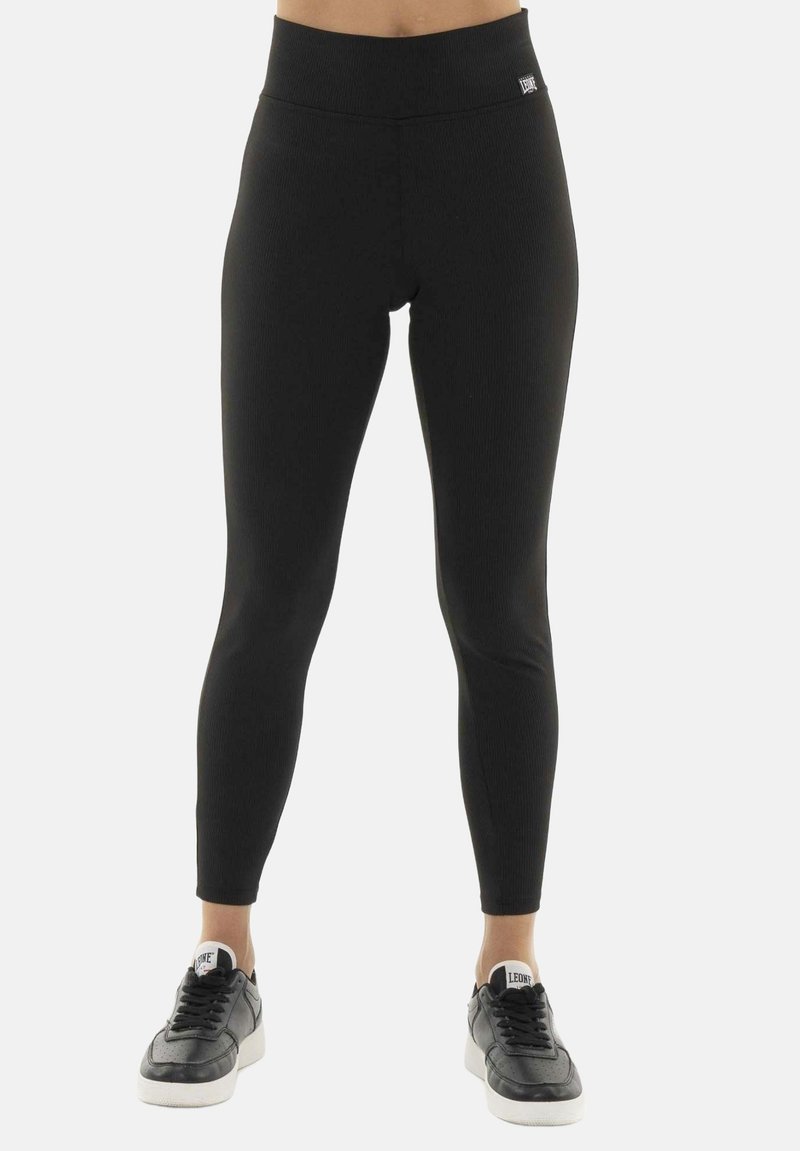 Personne portant des leggings noirs taille haute et des baskets noires avec semelles blanches, debout devant un fond clair uni.